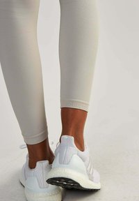 Leggings beige chiaro abbinati a scarpe da corsa bianche in rete, con suola testurizzata e accenti del marchio sul tallone e sulla linguetta.