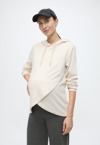 Vaaleanbeigeä äitiyscollegepaita, jossa on ristiin kietoutuva malli, säädettävä kiristysnauha ja pitkät hihat, yhdistetty tummanharmaisiin ribbineulottuihin housuihin.