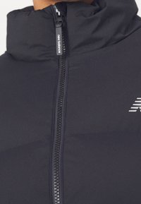 Zbliżenie na zamek błyskawiczny czarnej kurtki New Balance z wyściełanym kołnierzem i logo na piersi.