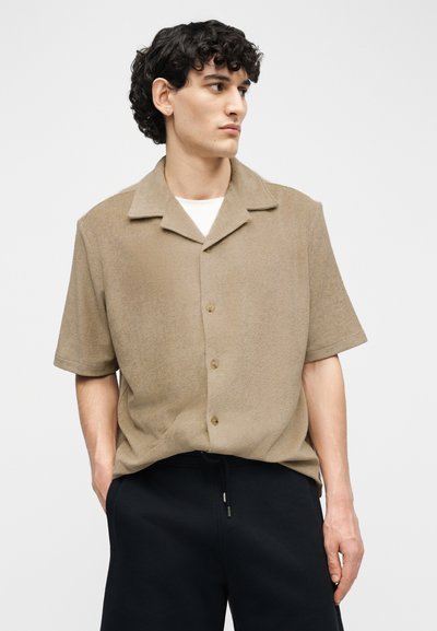 Νεαρός άνδρας με σγουρά μαύρα μαλλιά, φοράει μπεζ κοντομάνικο πουκάμισο με κουμπιά και μαύρο παντελόνι, κοιτάζοντας προς τα δεξιά.