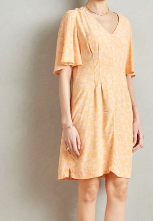 Personne portant une robe pêche à manches courtes avec un motif floral subtil et un décolleté en V, debout contre un mur clair uni.