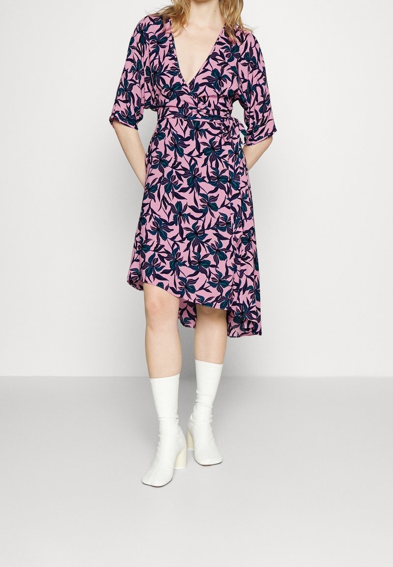 Robe portefeuille florale rose avec un motif de feuilles bleu foncé, décolleté en V et manches courtes, présentant un ourlet asymétrique. Portée avec des bottes blanches à talons.