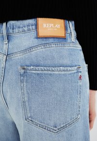 Svijetloplave traperice s teksturiranim završetkom, s kožnom etiketom s natpisom "REPLAY SINCE 1981" na struku i malim crvenim logom.