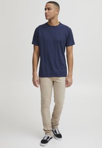 Temno modra bombažna majica s kratkimi rokavi in okroglim izrezom, v kombinaciji s svetlo bež slim-fit hlačami in črnimi supergami z belimi podplati.