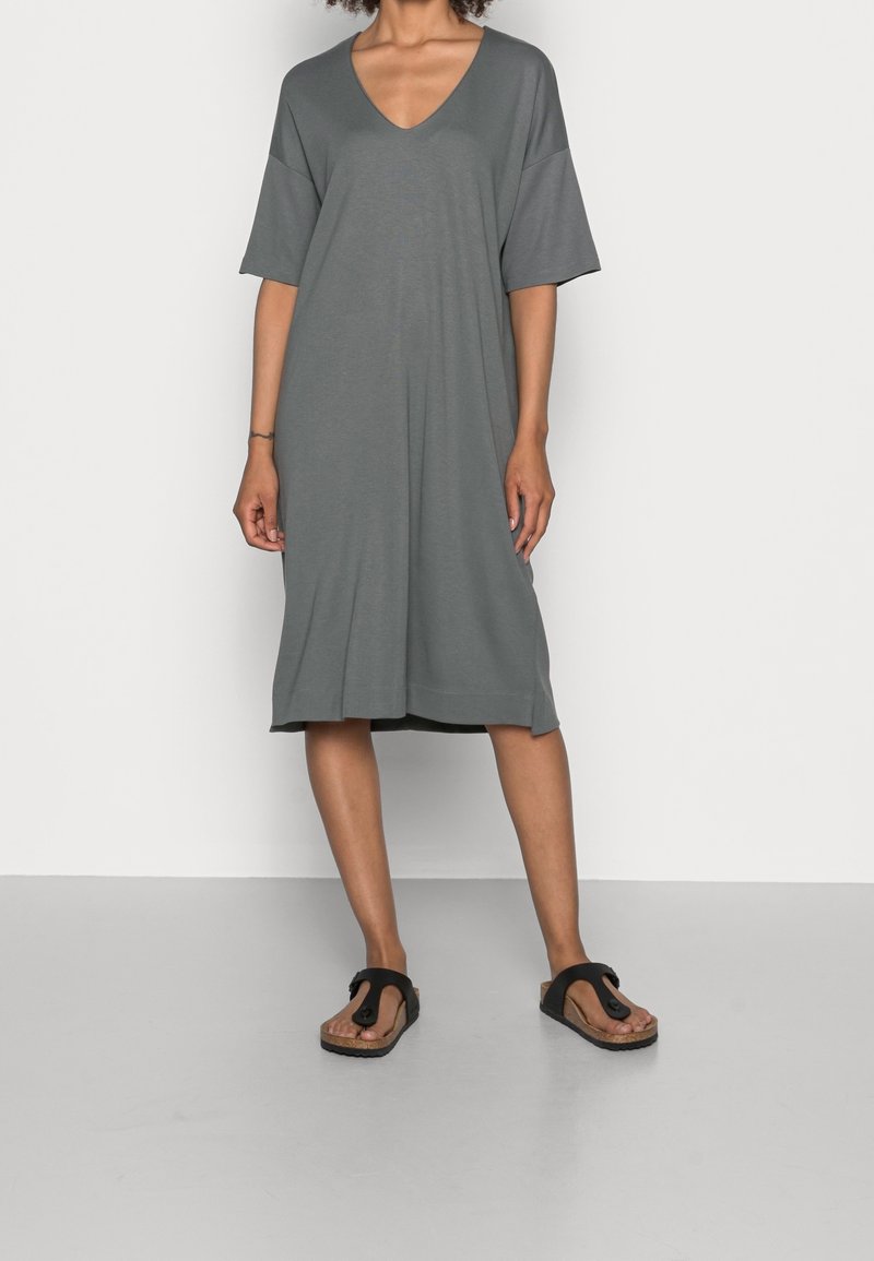 Femme portant une robe ample grise jusqu'aux genoux avec un col en V et des manches courtes, associée à des sandales noires à boucle sur les orteils, debout sur un sol gris.
