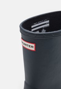 Botte de pluie en caoutchouc noire avec le logo "Hunter" blanc et rouge sur la tige supérieure, sur fond blanc.