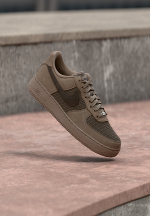 Hnedá teniska Nike Air Force 1 s detailmi z sieťoviny a kože levitujúca nad ružovým betónovým povrchom a sivým kamenným pozadím.