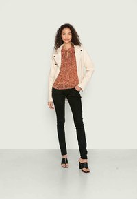 Jaqueta de couro sintético bege sobre uma blusa floral em cor ferrugem, jeans skinny pretos e sandálias pretas de salto robusto. Ambiente interior.