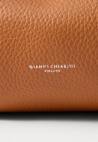 Iedegta tekstūra āda ar akmens virsmu. Baltā uzņēmuma logo "GIANNI CHIARINI FIRENZE" ir centrēts pa apakšējo malu.