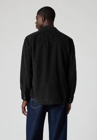 Camisa preta de veludo cotelê com pala pontiaguda, punhos com botões e bainha ligeiramente curva. Usada com jeans azul escuro. Material texturizado e resistente.