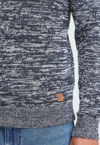 Artimiausias kadras, rodantis liemenį, vilkintį juodai-baltą margintą megztinį su ruduo logo etikete, derinamą su mėlynomis džinsomis.