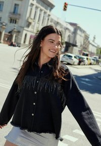 Juodas džinsinis švarkas su frakcinėmis detalėmis ir sidabriniais mygtukais, turintis trumpą dizainą ir susidėvėjusį kraštą. Fone matyti miesto gatvė.