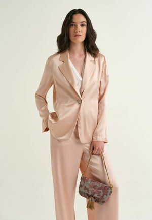 Donna che indossa un completo in raso beige con top bianco, tiene una borsa a tracolla decorata con nappine, in piedi davanti a uno sfondo chiaro e uniforme.