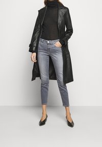 Femme portant un trench-coat en cuir noir, un col roulé noir, un jean gris court et des talons noirs, debout devant un fond blanc.