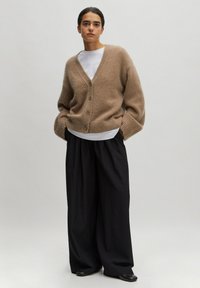 Beige wollen cardigan met V-hals en knoopsluiting, gecombineerd met losse zwarte broeken. Geverfde stof en relaxte pasvorm.