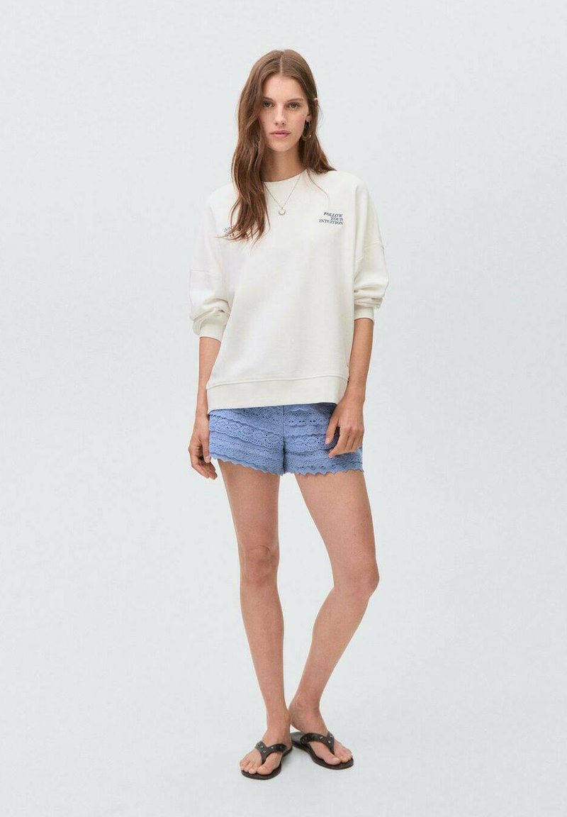 Light beige sweatshirt with cuffs, crew neck, and printed text. Wavy blue lace shorts with scalloped edges. Black flip-flops.