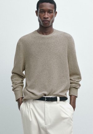 Giovane uomo che indossa un maglione beige di maglia e pantaloni bianchi con cintura nera, in piedi con le mani nelle tasche contro uno sfondo semplice.