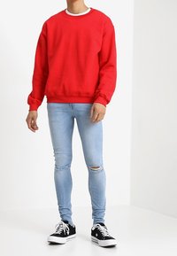 Mies, który ma na sobie czerwoną bluzę, jasnoniebieskie przetarte obcisłe dżinsy oraz czarne trampki z białymi sznurówkami, stojący na białym tle.