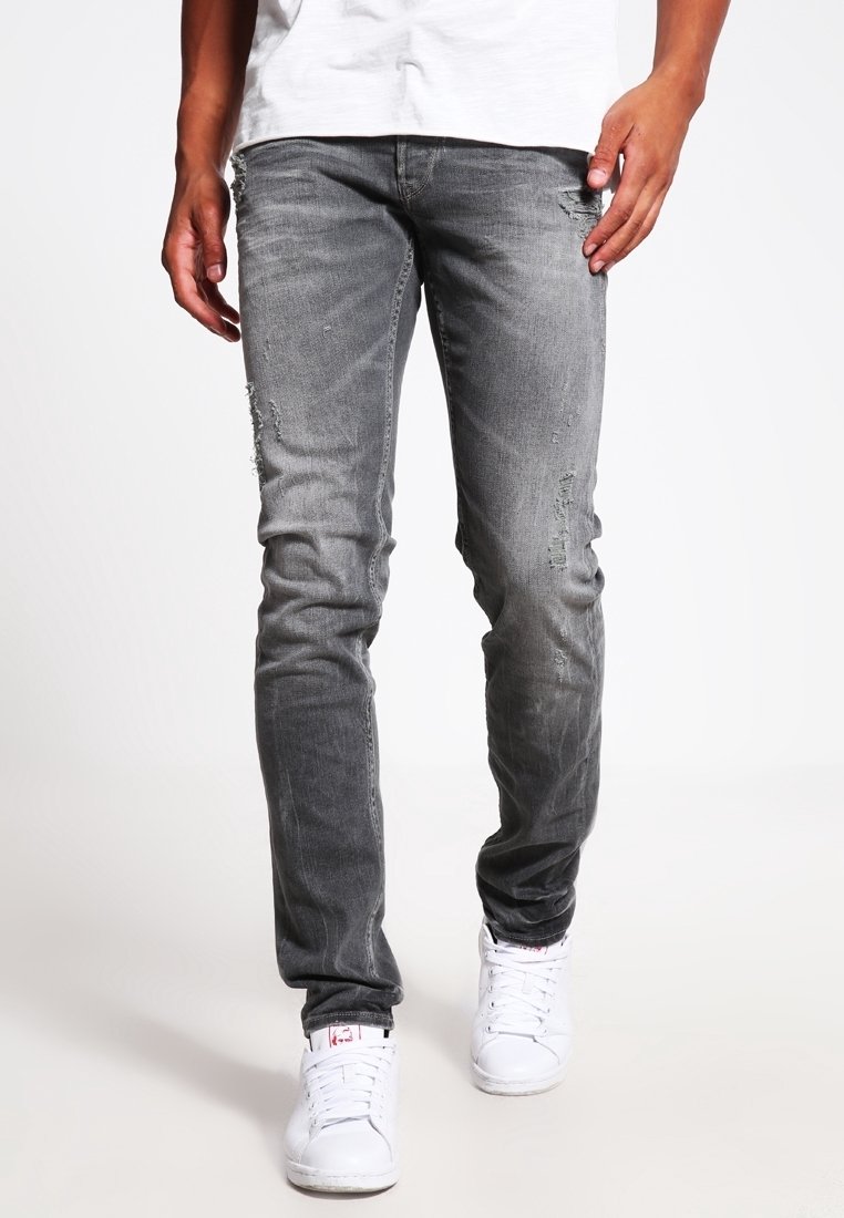 Jean gris slim déchiré porté avec des baskets blanches et un t-shirt blanc, visible de la mi-cuisse aux pieds sur un fond uni.