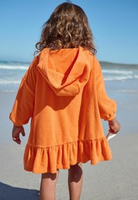 Pomarańczowy, kapturowy ręcznik plażowy wykonany z miękkiego materiału, z falbaniastym dołem i luźnymi rękawami, o gładkiej teksturze i swobodnym fasonie.