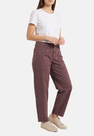 Mujer con camisa blanca de manga corta, pantalones marrones holgados con grandes bolsillos y zapatillas beige, de pie frente a un fondo liso.