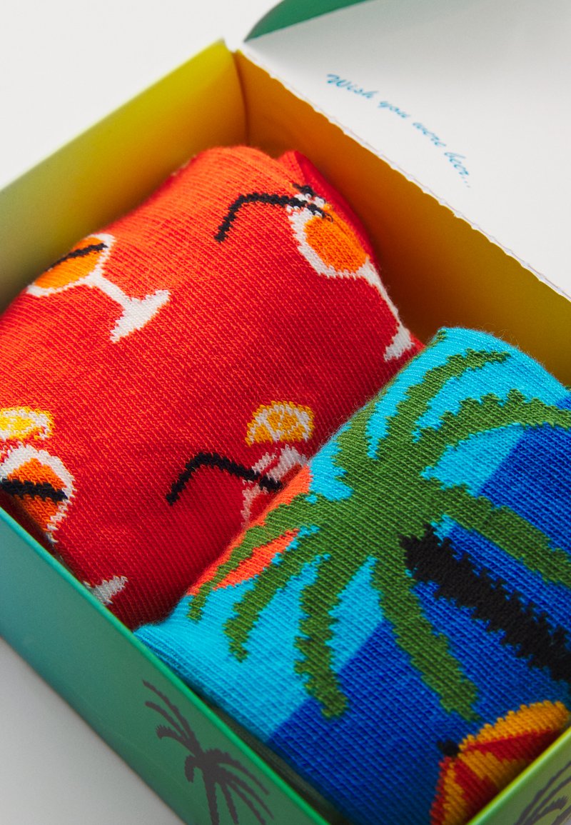 Zwei Paare bunter Socken in einer Schachtel: ein rotes Paar mit Cocktailmustern, das andere blaue mit Palmen, beide aus Strickstoff.