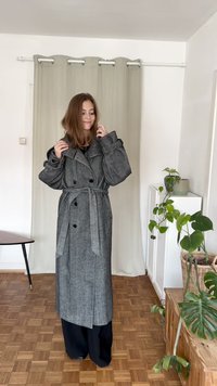 Trench coat cinza oversized com cintura com cinto, grandes lapelas e fechos de botão, usado sobre calças escuras, em frente a um fundo claro com plantas.
