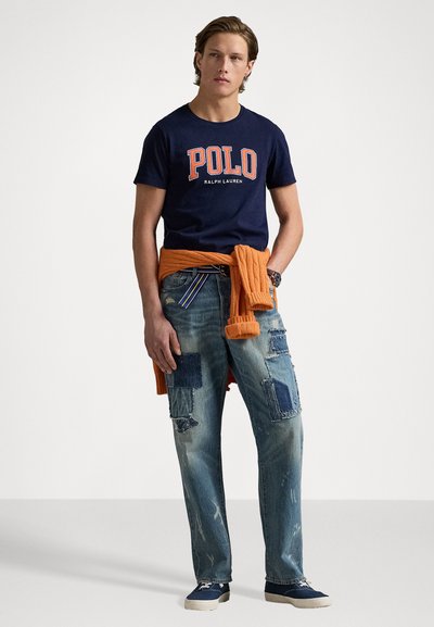 Tengerészkék póló "POLO" logóval, narancsszínű kötött pulóver a derékon megkötve, megviselt világoskék farmer, foltokkal, tengerészkék cipő.