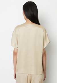 Blouse en satin beige à coupe décontractée, manches courtes et encolure ronde. Présente une texture lisse et un dos avec des coutures minimales.