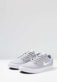 Baskets en toile grises avec semelles en caoutchouc blanches, présentant un design plat, un bout rond et le logo swoosh blanc de Nike sur le côté.