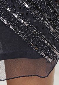 Svart nät tyg med paljetter i rader, med en genomskinlig nederkant. Glittrande detaljer skapar struktur och visuell intress.