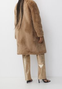 Manteau en fausse fourrure marron à manches longues et coupe décontractée, associé à un pantalon métallique doré brillant et des sandales noires à talons hauts.