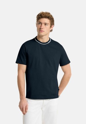 Giovane uomo con capelli ricci che indossa una camicia nera a maniche corte con bordo bianco sul colletto e pantaloni bianchi, che guarda di lato.