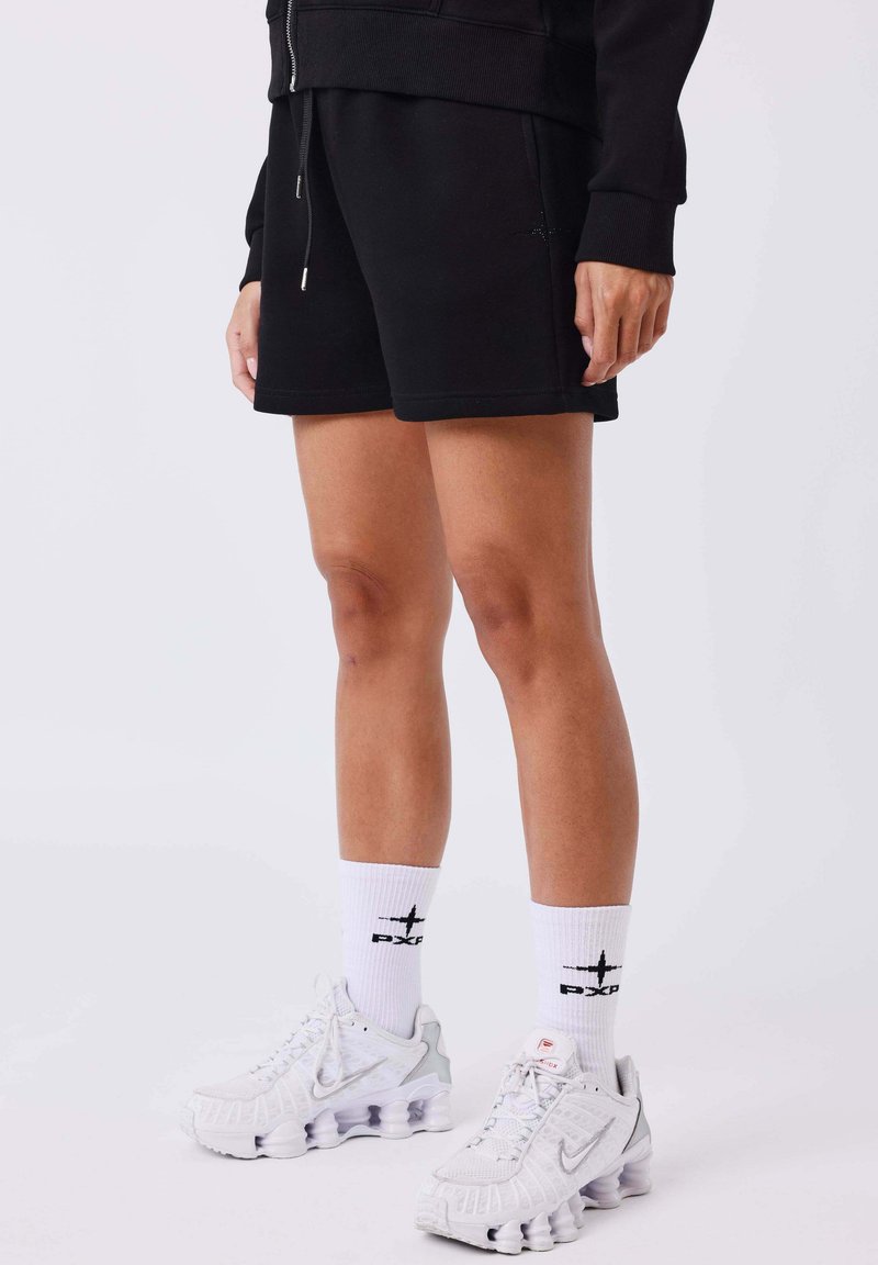 Personne portant un short noir, un haut noir zippé, des chaussettes blanches montantes avec un logo noir, et des chaussures de course blanches Nike, debout sur un sol blanc.