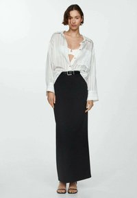 Vrouw staat gekleed in een witte gestreepte losse blouse over een witte bralette, gecombineerd met een hoog-waist zwarte maxi rok en zwarte sandalen.