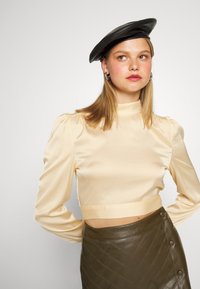 Top cropped de satin creme com mangas longas, decote alto e ombros franzidos, combinado com uma saia de couro verde escuro com padrão acolchoado.