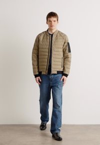 Casaco bomber em khaki acolchoado com punhos e bainha em malha preta, apresentando um bolso lateral. Usado com jeans azuis e sapatos pretos.