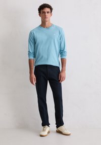 Lichtblauw shirt met lange mouwen en een ronde hals, gecombineerd met donkerblauwe broek en beige sneakers, tegen een neutrale achtergrond.