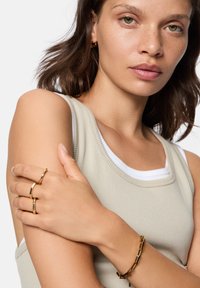Beige geribbelde tanktop over een witte onderhemd; het model draagt gouden ringen en een dikke gouden kettingarmband om haar pols.
