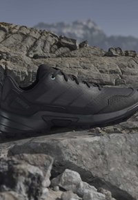 Black outdoor trail shoe with textured sole and laces resting on rocky terrain with blurred mountainous background.
