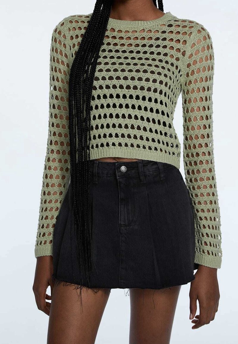 Maglione verde lavorato a maglia con design ad aperture, maniche lunghe e orlo cortissimo, abbinato a una gonna di jeans nera con orlo sfrangiato.