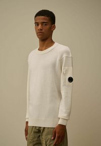 Maglione bianco lavorato a maglia con polsini e orlo a costine, dotato di una tasca a toppa con un accento circolare nero sulla manica sinistra. Taglio casual e testurizzato.
