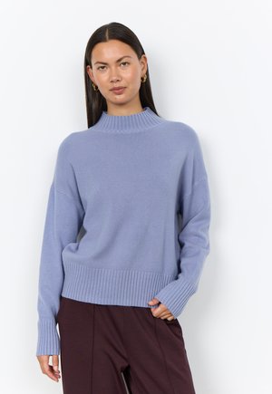 Woman wearing light blue ribbed high-neck sweater and dark brown pants, standing against a plain white background.