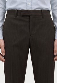 Homme portant un pantalon de costume marron foncé à fines rayures et une chemise bleu clair boutonnée, les mains détendues sur les côtés.