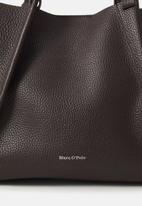Sac fourre-tout en cuir brun foncé avec une surface texturée, arborant le nom de la marque "Marc O'Polo" en blanc en bas.