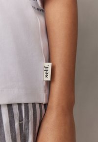 Bijela pamučna majica s tiskanim tekstom, s malom tkanom etiketom sa strane na kojoj piše "self." Ispod se vidi sivi prugasti materijal.