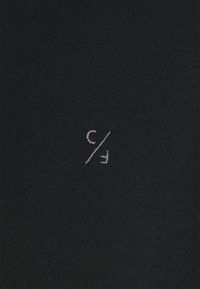 Schwarzer gestrickter Stoff mit einem kleinen, weißen gestickten Logo, das ein "c" und ein " / "-Symbol zeigt, das zentral auf dem Material platziert ist.