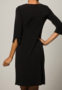 Schwarzes knielanges Kleid mit Dreiviertelärmel und Schlitzdetails, in lässiger Passform, aus glattem Stoff mit subtiler Glanzwirkung.