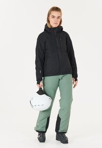 Schwarze wasserdichte Jacke mit Kapuze, kombiniert mit grünen Schneehosen mit schwarzen Akzenten. Die Person hält einen weißen Skihelm in der Hand.
