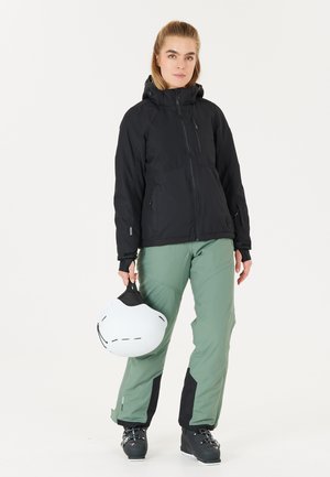 Black waterproof jacket with a hood, paired with green snow pants featuring black accents. The person holds a white ski helmet.